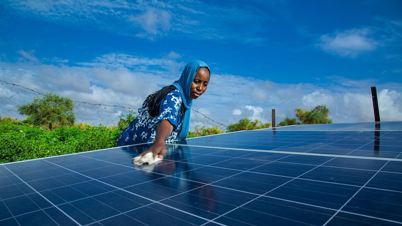 A photo of a woman cleaning a solar panel illustrates an op-ed on gender-smart climate investing
